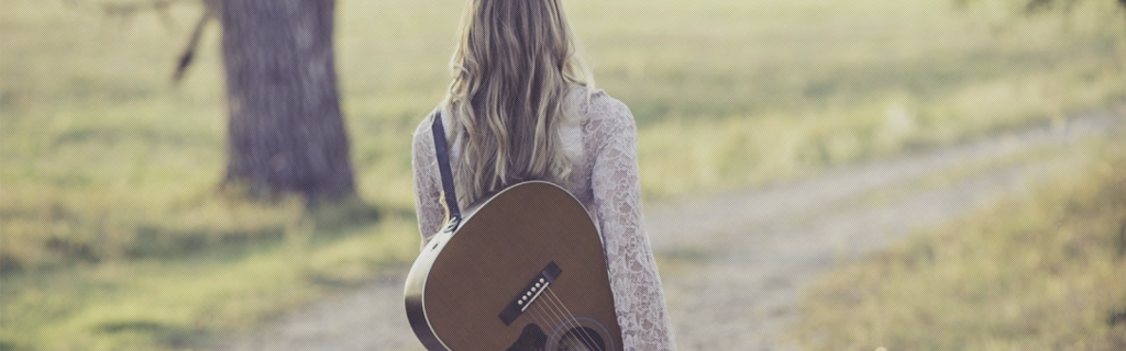 Frau mit Gitarre über der Schulter geht über ein Feld spazieren, man sieht sie von hinten